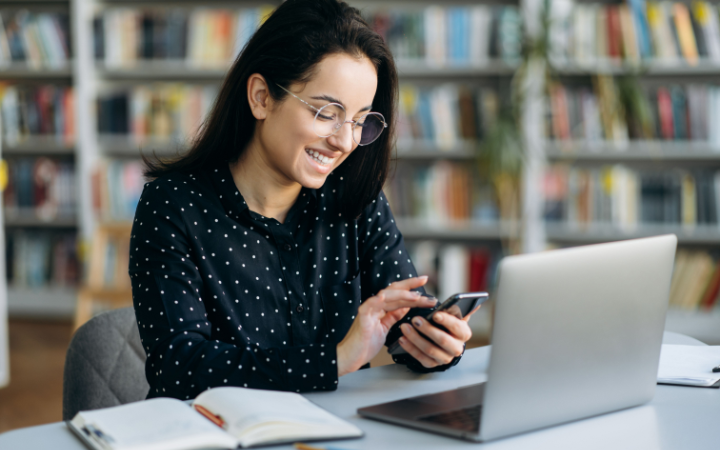 Eine Frau schaut auf ihr Smartphone und lacht. Vor ihr steht ein Laptop auf dem Schreibtisch.
