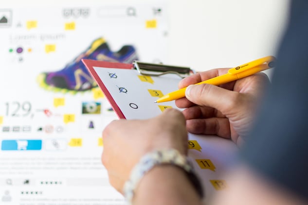 Jemand hält ein Clippboard in der Hand und kreuzt dort verschiedene Kategorien an, während im Hintergrund die Produktseite eines Online-Shops zu sehen ist.